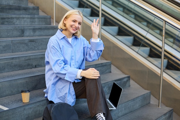 Free photo friendly student girl saying hi waving hand at someone sitting on stairs and drinking coffee using