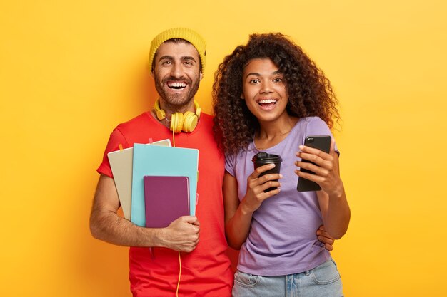 friendly stylish couple posing against the yellow wall with gadgets