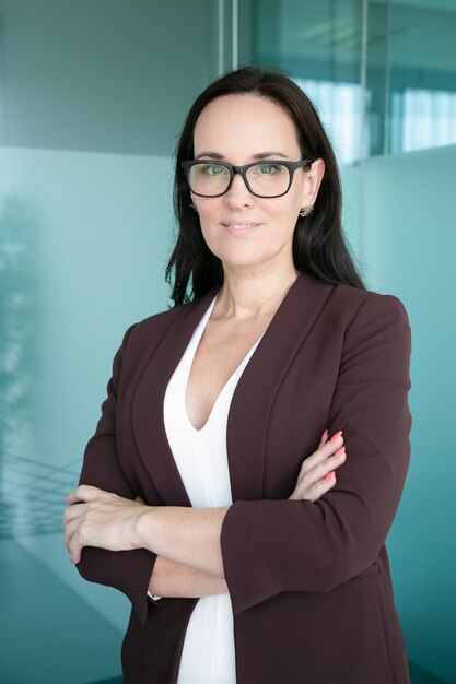 Friendly successful business lady wearing formal suit and glasses, standing with arms folded  and smiling