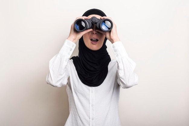 白い​シャツ​と​ヒジャーブ​の​フレンドリー​な​驚いた​若い​イスラム​教徒​の​女性​は​双眼鏡​を通して​見る​バナー​は​カメラ​を​見る