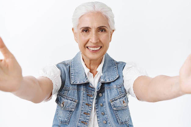 Free photo friendly tender and kind senior woman with grey combed hair reaching hands forward hold camera as taking selfie photographing or holding someone to give hug cuddle you with smile