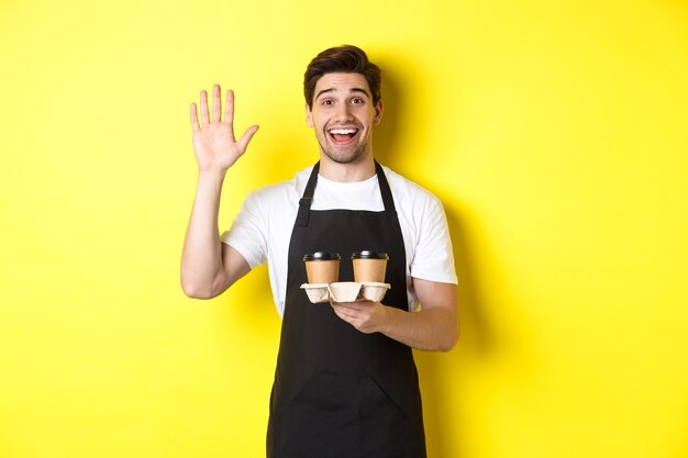 Friendly waiter in cafe waving hand at customer, holding takeaway coffee oder, standing against yellow background in black apron.