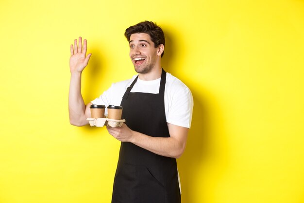 Friendly waiter in cafe waving hand at customer holding takeaway coffee oder standing against yellow...
