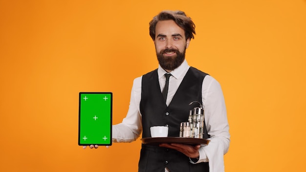 Friendly waiter shows tablet with greenscreen on camera, presenting blank copyspace mockup template. experienced butler in suit holding device with isolated empty display and food tray