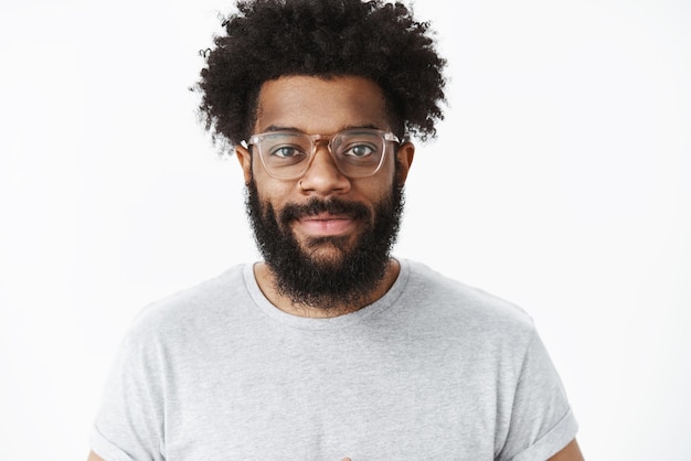 Дружелюбно выглядящий общительный и симпатичный афроамериканский бородатый мужчина цифровой кочевник в очках с афро-прической улыбается с восторженным и напористым выражением лица, позируя над серой стеной
