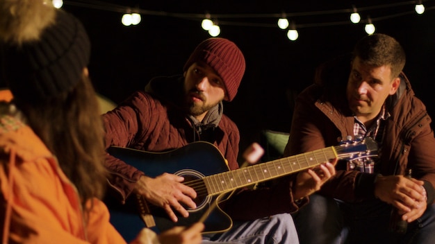 Friends engjoying a guitar solo from one their friends in camping. Cold night in autumn. Retro camper van.