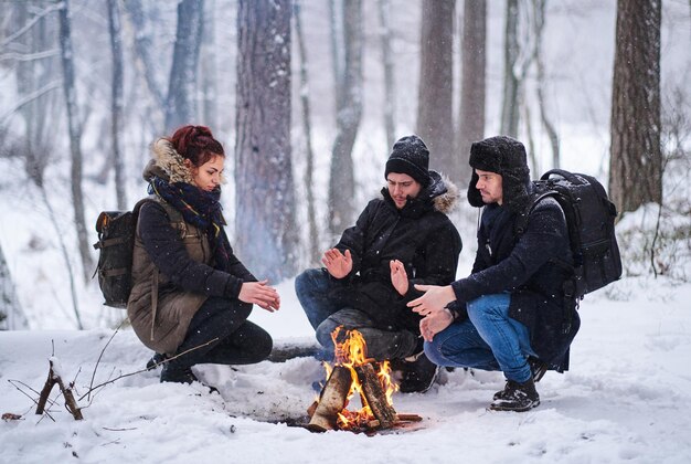 Friends hike in the snowy forest. Young hikers heated by the fire.