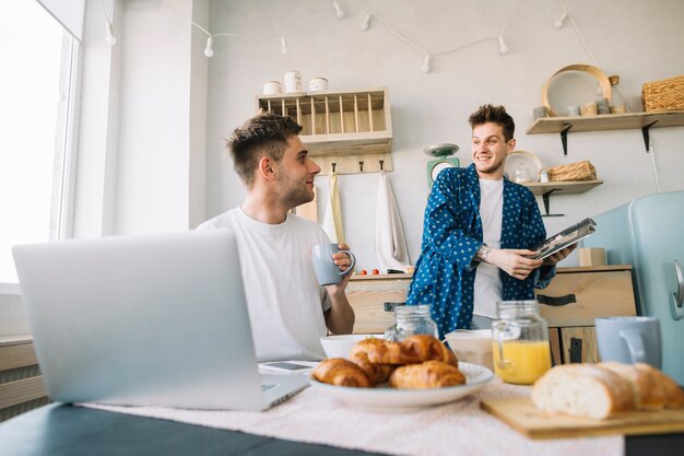 Friends looking at each other holding magazine and cup of coffee in kitchen
