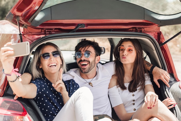 Foto gratuita amici che si siedono nel bagagliaio dell'auto prendendo selfie tramite smartphone