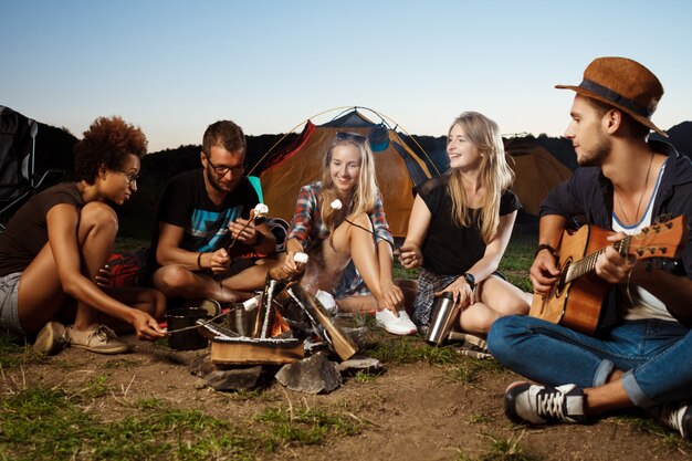 Friends sitting near bonfire, smiling, playing guitar