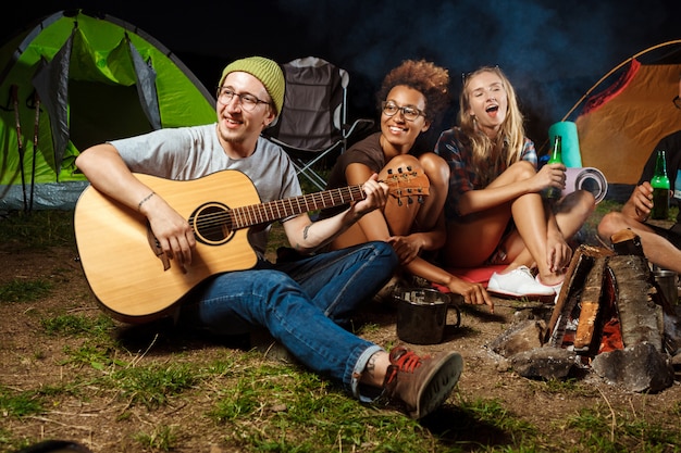 Friends sitting near bonfire, smiling, speaking, resting, playing guitar