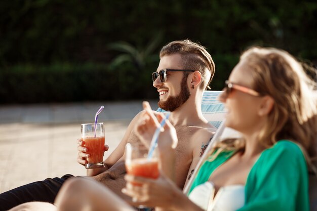 Friends smiling, drinking cocktails, lying on chaises near swimming pool