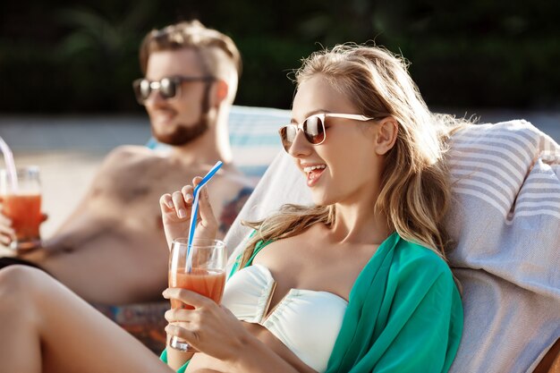 Friends smiling, drinking cocktails, lying on chaises near swimming pool