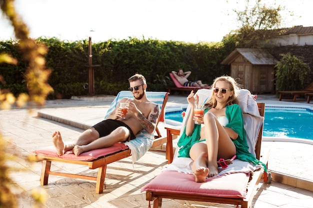 Friends smiling, drinking cocktails, lying on chaises near swimming pool