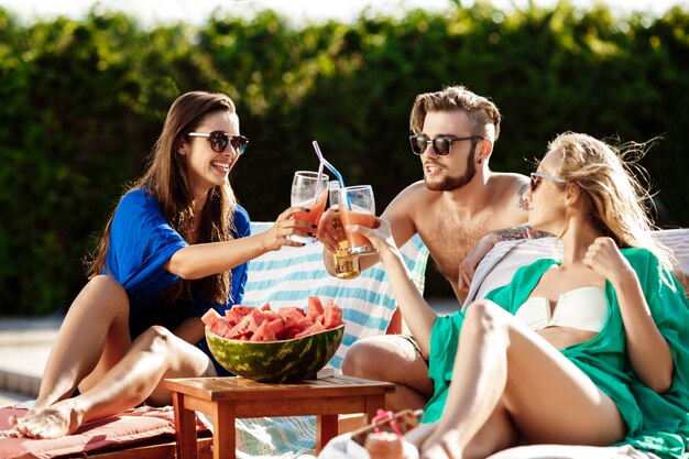 Friends smiling, eating watermelon, drinking cocktails, relaxing near swimming pool