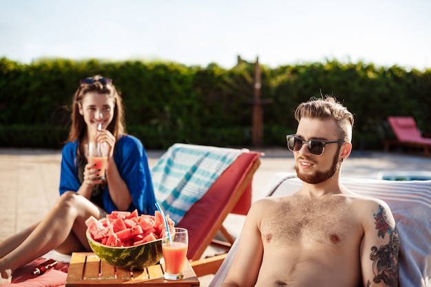 Friends smiling, resting, drinking cocktails, lying near swimming pool