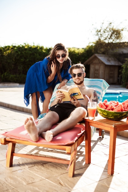 Friends sunbathing, reading book, lying near swimming pool