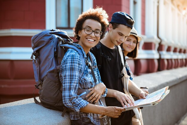 Friends travelers with backpacks smiling, looking route at map in the street.