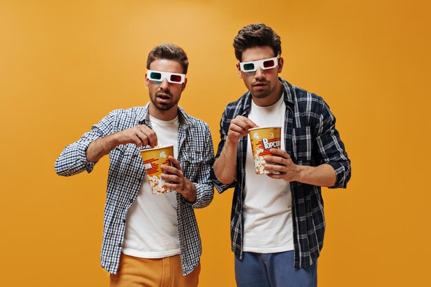 Friends watch movie in 3D eyeglasses on orange background Brunet men in checkered shirts eat popcorn and pose on isolated