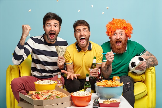 Friendship, game, gambling, leisure concept. Emotional excited three male friends watch football game on tv at home, clench fists, shout during goal