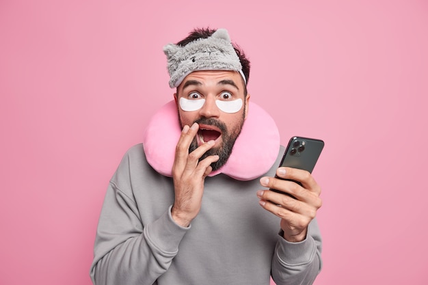 Испуганный бородатый взрослый мужчина выглядит напуганным, держит современный смартфон, носит маску для сна, удобную подушку на шее