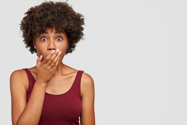 Foto gratuita spaventata bella donna dalla pelle scura con taglio di capelli afro, copre la bocca e fissa, vestita con una t-shirt casual, reagisce alle fake news, sta contro il muro bianco