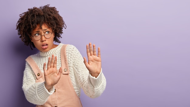 Free photo frightened dark skinned female outstretches palms, makes protective gesture, has worried nervous facial expression, asks not come closer, wears optical glasses and overalls, isolated on purple wall