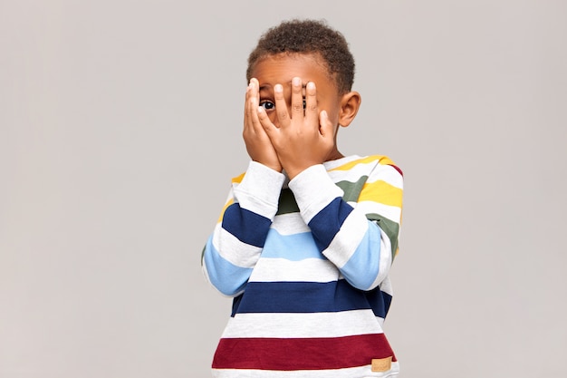 Frightened dark skinned little boy covering face with both hands as if being afraid to see something scary, spying through hole between fingers. Shy African child hiding, or playing hide and seek