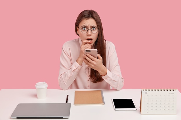 frightened displeased woman holds mobile phone, has everything neatly arranged on table