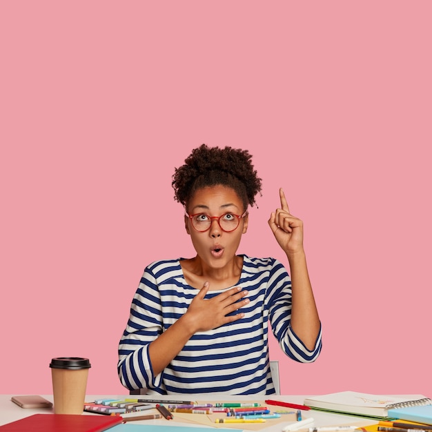 Free photo frightened emotional dark skinned female designer raises index finger, keeps hand on breast, points upwards, notices unbelievable item, uses notebook, caryons for making sketch sits in coworking space