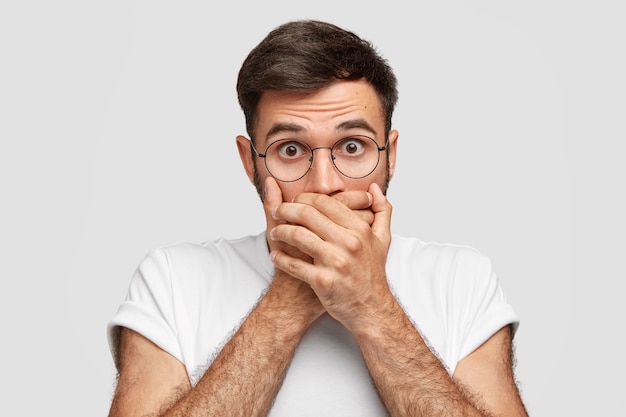 Free photo frightened handsome man closes mouth with both palms, stares with bugged eyes