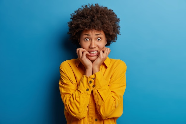 Frightened nervous woman clenches teeth, panicking as being in danger, stands embarrassed, wears yellow shirt, isolated over blue background.