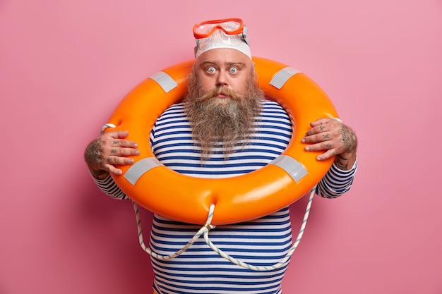 Frightened overweight holidaymaker afraids of sinking, uses safety equipment, wears snorkeling goggles, swims with lifebuoy, looks directly with shocked expression. travel insurance