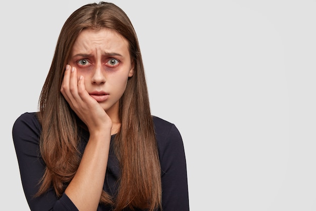 Frightened young woman with bruises posing against the white wall