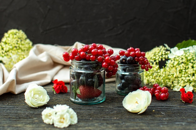 Free photo a front close up view fresh berries such as blackberries and blueberries inside glass cans around white flowers on the grey wooden floor