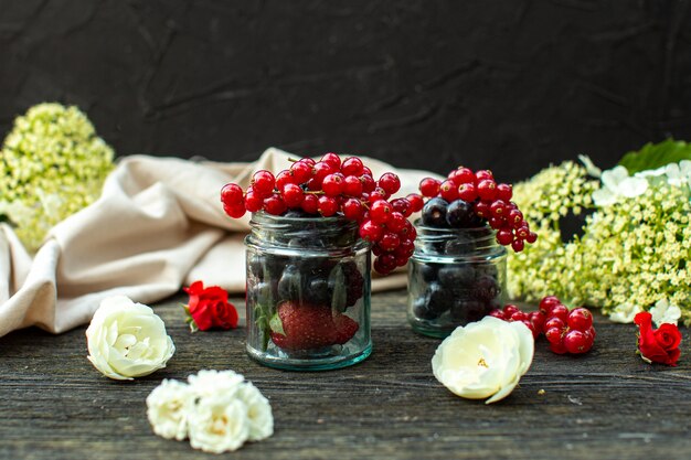 A front close up view fresh berries such as blackberries and blueberries inside glass cans around white flowers on the grey wooden floor