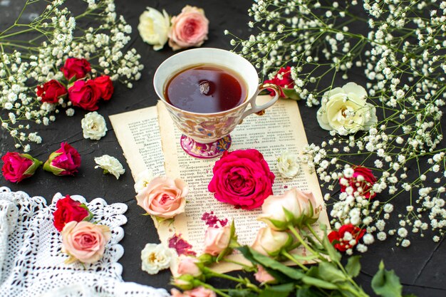 A front close up view hot tea on the paper and around different colored roses on the grey surface