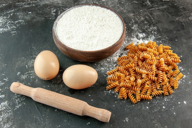 Front close up view of raw Italian pastas two eggs and flour in a brown bowl rolling-pin on gray background