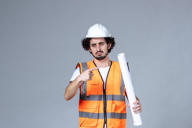 Front close view of angry male construction worker in warning vest with safety helmet and holding blank on gray wall