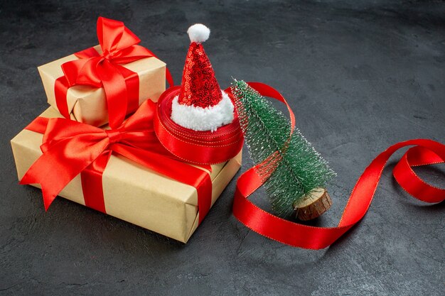 Front close view of beautiful gifts with red ribbon and santa claus hat christmas tree on dark table