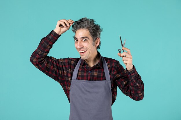 Front close view of confident male hairdesser wearing gray apron and holding scissor combing his hair on blue soft color background