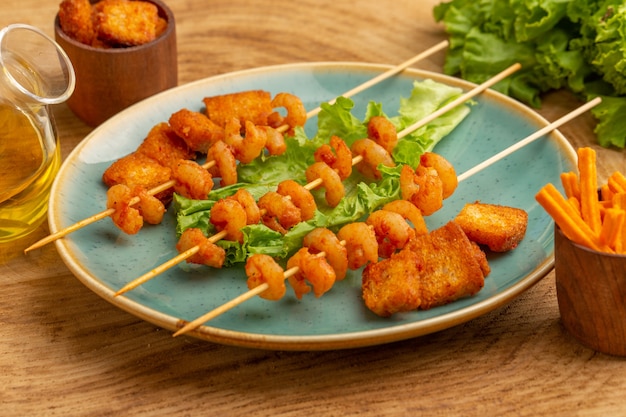 Front close view cooked shrimps on sticks inside blue plate with green salad and oil on wood