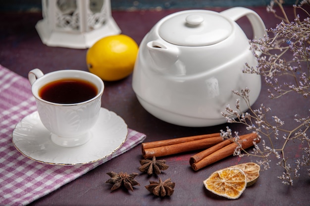 Front close view cup of tea with cinnamon and kettle on dark surface tea drink lemon color