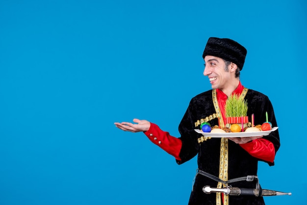 Front close view of curious happy young guy in traditional dress holding tray filled with national confectionery and pointing something on the right side on blue background