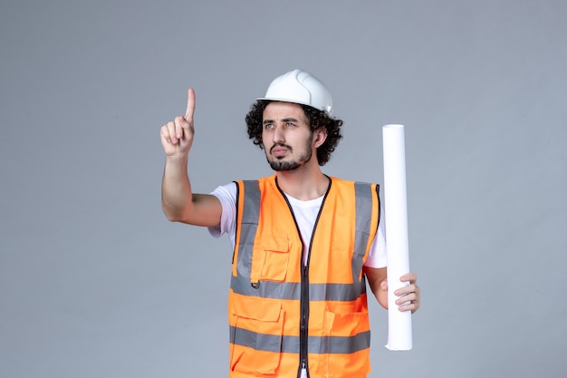 Front close view of curious male construction worker in warning vest with safety helmet and showing blank pointing up on gray wall