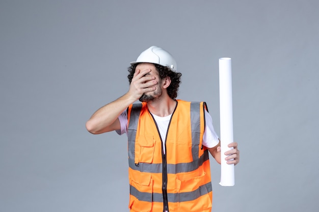 Front close view of exhausted male construction worker in warning vest with safety helmet and holding blank on gray wall