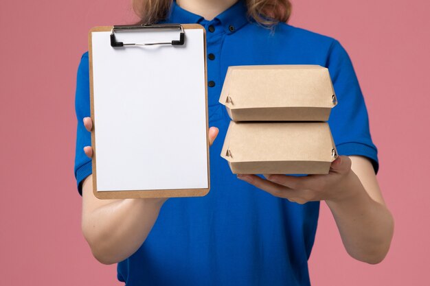 Front close view female courier in blue uniform cape holding little delivery food packages and notepad on the pink desk delivery service job employee