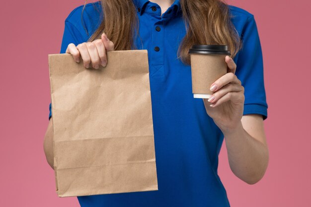 Крупным планом вид спереди женщина-курьер в синей форме, держащая коричневую кофейную чашку с пакетом продуктов на светло-розовом столе, служба доставки униформы