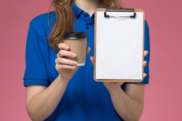 Front close view female courier in blue uniform holding brown coffee cup with notepad on the light-pink desk service job uniform delivering company