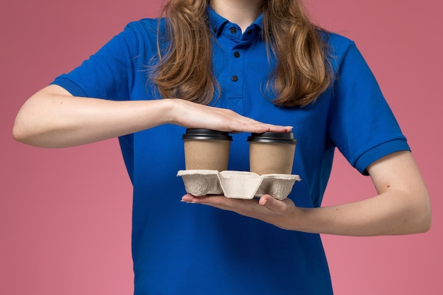 Foto gratuita corriere femminile di vista ravvicinata anteriore in uniforme blu che tiene le tazze di caffè di consegna marrone sul lavoro dell'azienda uniforme di servizio del fondo rosa